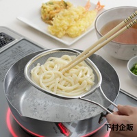 一只手正握著帶有麵條的不鏽鋼橢圓形濾網，濾網浸在裝有滾水的不鏽鋼鍋中。一雙木筷子夾著麵條，置於濾網上方。背景是裝有炸物的盤子和一碗湯。