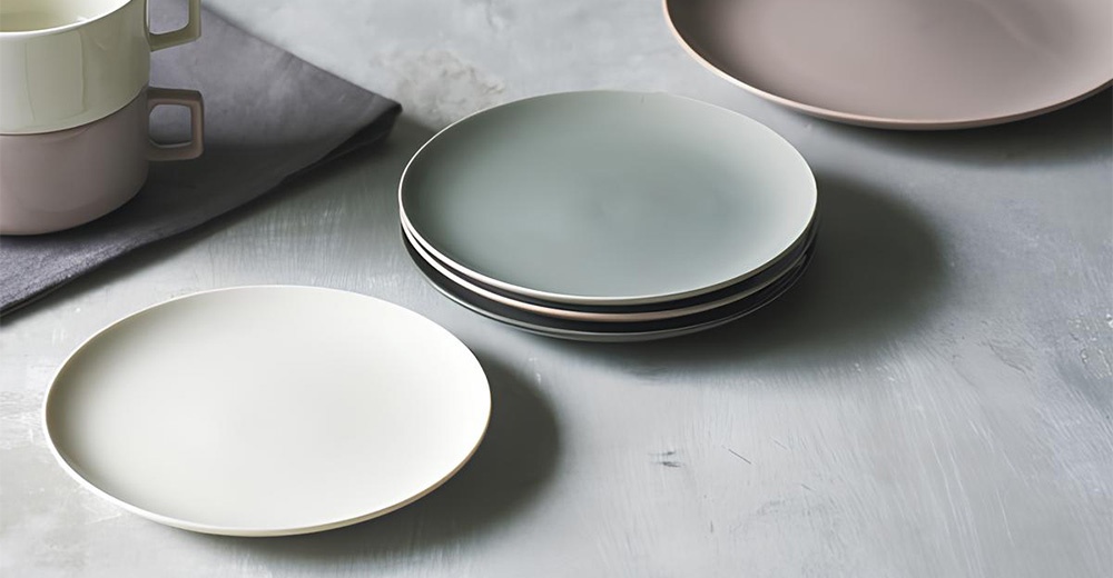 Aito 美濃燒簡約素雅質感圓餐盤6入組，包含單個白色圓盤、疊放的灰色圓盤組，以及上方有疊放的淺綠色和粉色馬克杯，皆擺設於灰色水泥紋理的桌面。