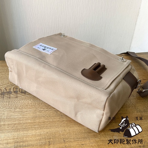 米色純棉帆布製成的橫式側背包，配有咖啡色皮革裝飾和金屬扣環，包蓋上印有品牌標籤。
