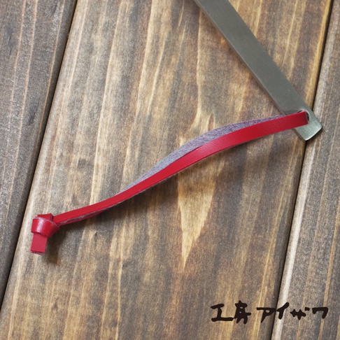 不鏽鋼萬用湯勺，勺柄處有紅色皮革和灰色絨布裝飾，皮革末端打結，背景為木紋桌面。