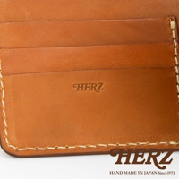 職人手作皮革二折橫式卡片紙鈔小短夾，近距離特寫，展現溫潤的棕色皮革材質、多個卡片夾層，以及壓印的「HERZ」品牌標誌和縫線細節。