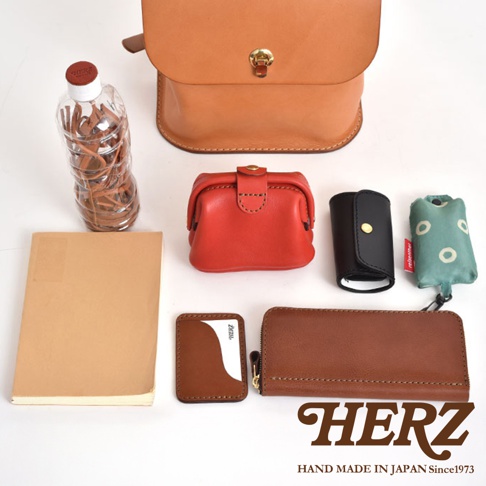職人手作皮革翻蓋迷你後背包，搭配紅色零錢包、黑色鑰匙包、綠色環保袋、棕色卡夾、長夾及一本筆記本，與一瓶裝滿皮革製品的透明瓶子。