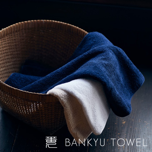 藤編籃中疊放著三條毛巾，顏色分別為深藍色、米白色和淺棕色。毛巾材質蓬鬆，質感柔軟。