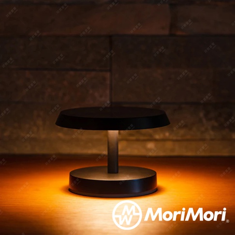 黑色金屬材質的LED T-Light LOUNGE工型造型檯燈，圓形燈罩下有簡潔的燈柱連接圓形底座，散發溫暖的黃色光芒，照亮桌面。