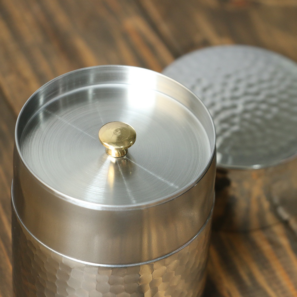 不鏽鋼錘目紋茶葉罐，頂部有金色圓形把手，罐身帶有手工錘擊紋路，質感細緻，光澤明亮。