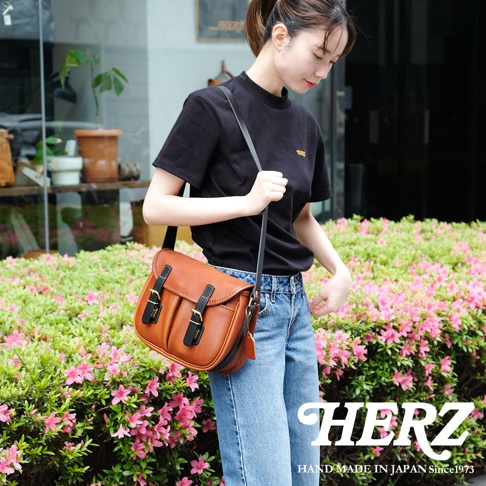 一位女性身穿黑色T恤和牛仔褲，肩背一個棕色皮革製的復古釣魚包，包上有兩個黑色搭扣，包身線條流暢，充滿質感。