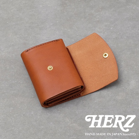 一款採用棕色Minerva Box皮革製成的日本HERZ職人手作Organ迷你短夾，錢包呈半展開狀態，展示了簡約的折疊結構與金色金屬按扣細節，整體呈現出溫潤的皮革質感與精緻的手作工藝。