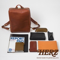 職人手作皮革軟皮商務後背包，搭配同款棕色皮革筆袋、黑色皮夾、黑色皮質平板電腦包、棕色皮革筆記本、橘色皮革文件夾，以及一份標示「REPORT PAD」的報告。