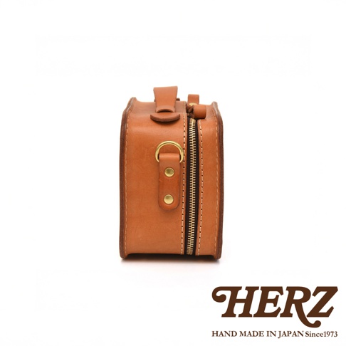 HERZ 品牌標誌，下方有 HAND MADE IN JAPAN Since1973 字樣，圖片中間是一個以皮革製作的棕色小包，包包側面有拉鍊和金屬環。