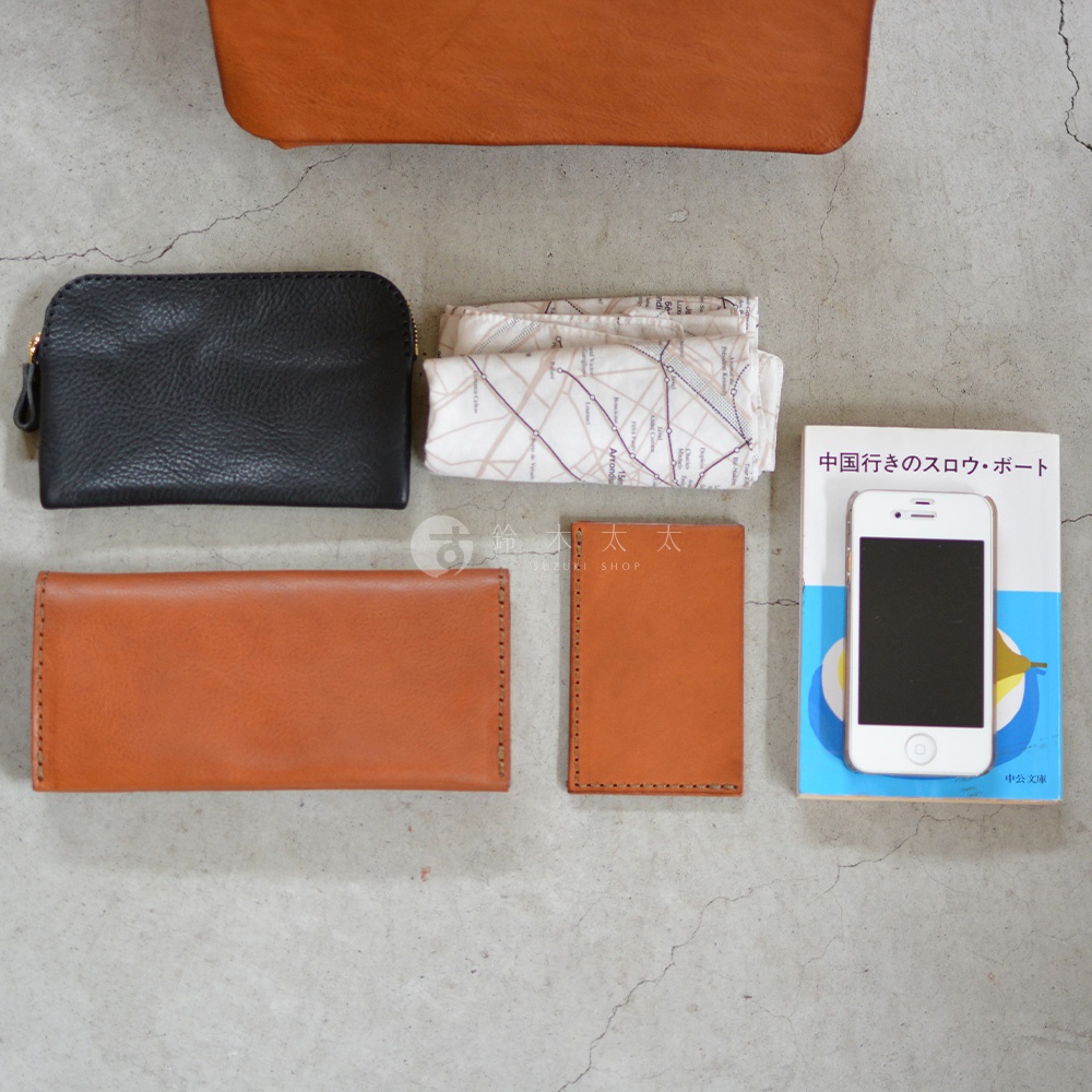 從上方視角看，一個黑色皮革化妝包、一個印有地圖圖案的白色布料、一個長款棕色皮革錢包、一個方形棕色皮革小冊子、一部白色手機及一本藍色封面的書，整齊擺放在灰色的水泥地面上。