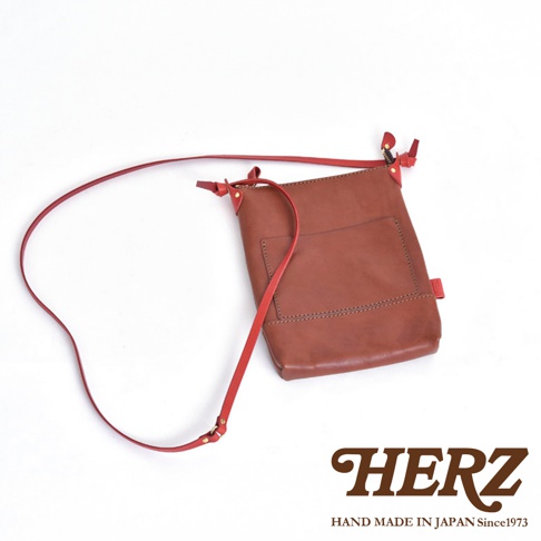 棕褐色皮革直式隨身單肩包，配有紅色皮革肩帶和細節，包身有縫線裝飾和一個外部口袋。