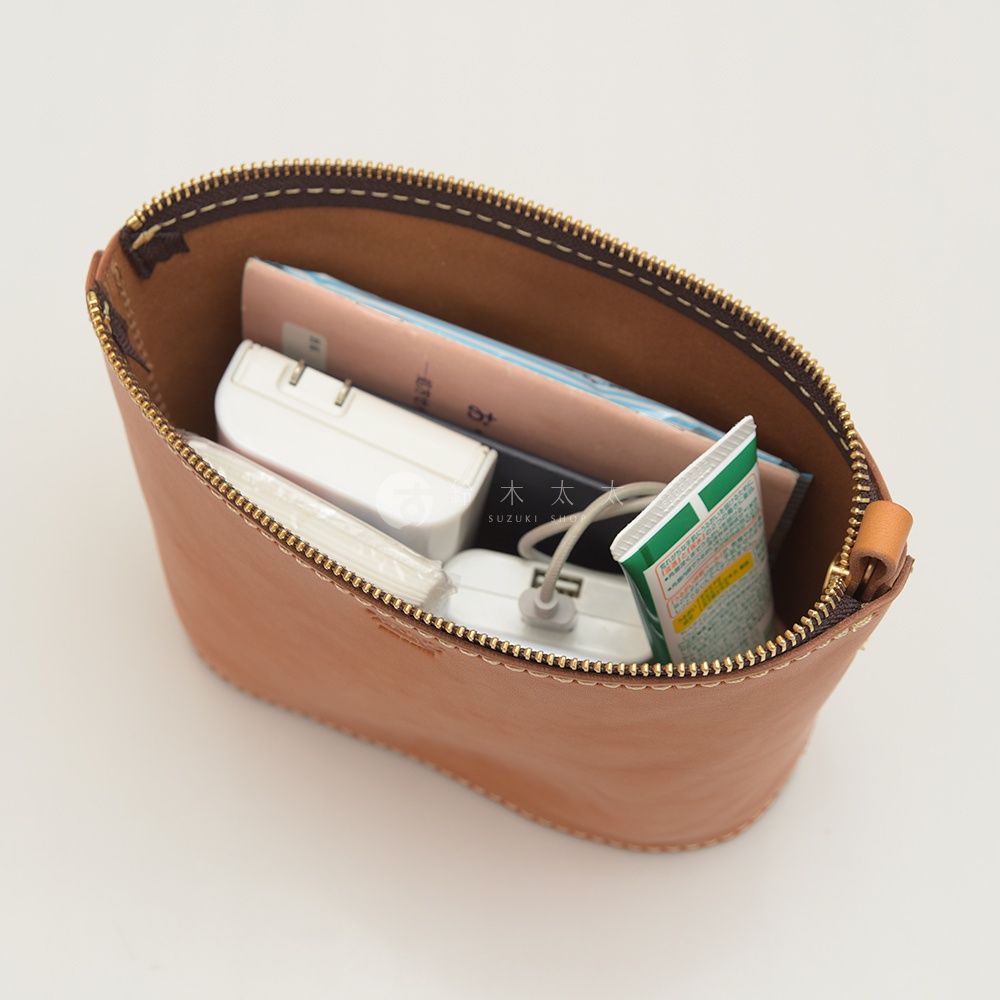 職人手作皮革三角收納包，淺棕色牛皮材質，內含一本書、白色行動電源、牙膏管狀物，拉鍊為金色。