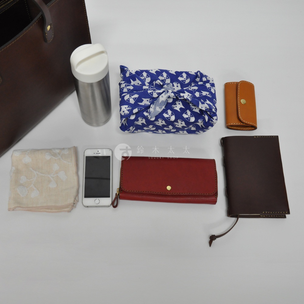 深棕色皮革直式托特包，內含一個銀色保溫瓶、藍色底白色貓咪圖案的布包、一個棕色皮革鑰匙包、一個米白色繡花手帕、一支白色手機、一個紅色長夾和一本深棕色筆記本。