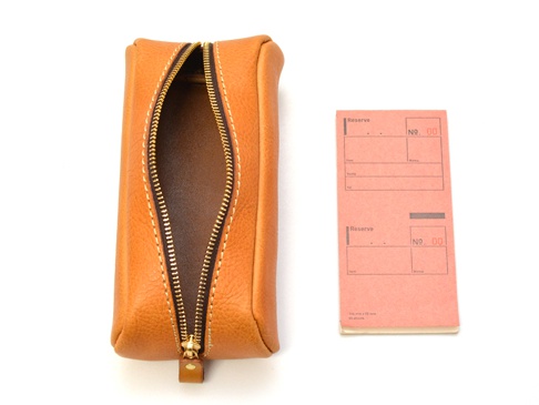 職人手作皮革圓角收納筆袋，淺棕色皮革材質，拉鍊半開，露出內部深棕色皮革。旁邊有一個粉紅色小筆記本，印有英文 Reserve 和數字 No. 00。