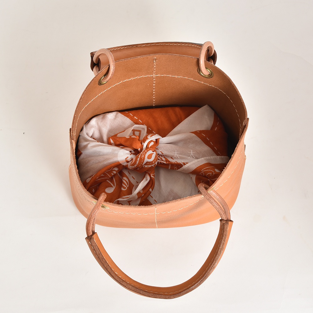 淺棕色皮革製成的大開口午餐袋，內有橘白圖案的包袱，提把為皮革材質，附有車縫線裝飾。