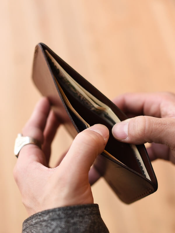 一只手拿著咖啡色皮革紙鈔夾，裡面放著紙鈔。另一隻手正在將紙鈔放入夾中。皮革表面光滑，夾子設計簡潔。