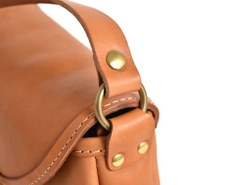 職人手作的淺棕色皮革馬鞍包，設有圓形金屬環及黃銅铆釘，搭配縫線細節，展現經典休閒風格。