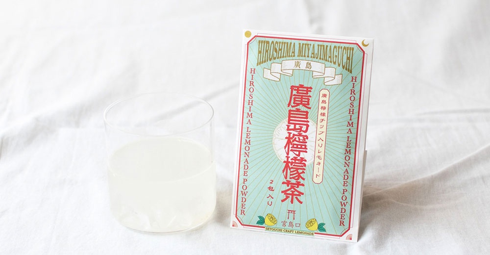 一張放置在白色皺褶布料上的廣島檸檬茶粉包裝盒，包裝採淺綠色與紅色配色，正中央以大字體寫著廣島檸檬茶，下方標註兩包入及宮島口字樣，包裝左側放著一杯已沖泡好的淡白色檸檬飲料，整體呈現清新雅致的氛圍。