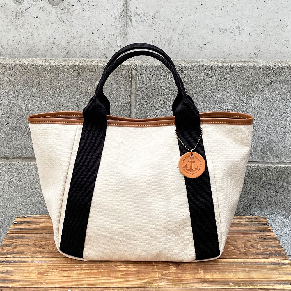 高島帆布多口袋手提托特包，採用米白色帆布材質，搭配黑色帆布提把與皮革滾邊。包包正面有黑色條紋裝飾，並掛有船錨圖案的皮革吊飾。