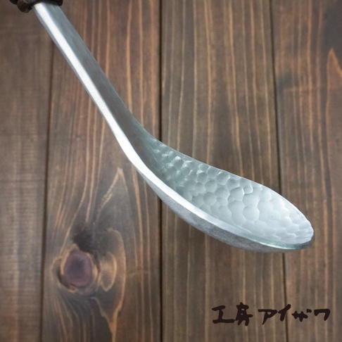 一把銀色鋁製湯勺，勺面內側有錘目紋，勺柄末端為深色木質。