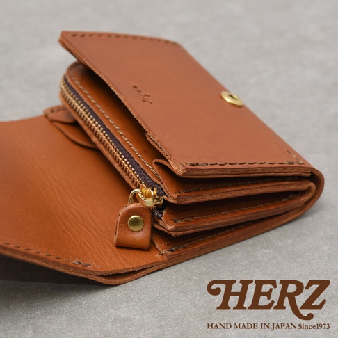 一張由日本品牌HERZ手工製作的棕色皮革雙折短夾特寫，呈現極簡俐落的設計風格，採用義大利Minerva Liscio皮革製作，表面保留細緻天然紋路，右下方印有品牌標誌與手工產地字樣，內部配備拉鍊式零錢袋及多層收納隔間，展示了細膩的手縫工藝與溫潤的皮質質感。