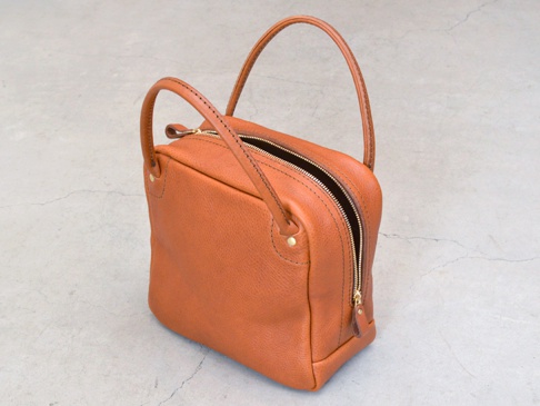 橘棕色皮革方形托特包，頂部有拉鍊開合，配有短提把，包身縫線細緻，側面有金色卯釘裝飾。