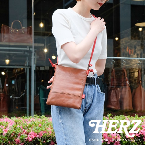 咖啡色皮革直式隨身單肩包，配有紅色細肩帶，包身有縫線裝飾和外部口袋。