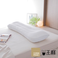 白色網狀材質的長條形枕頭，具有特殊曲線設計，放置於白色床單上，背景有植物裝飾與暖色圓形檯燈。