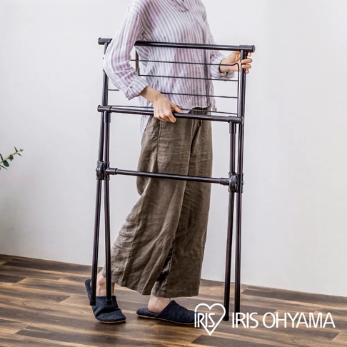一位穿著條紋上衣與寬褲的女性，正雙手拿著一台深棕色的 IRIS OHYAMA 室內折疊式曬衣架，展示其輕便收納的特性，曬衣架呈現長方形框架結構，設有水平橫桿方便掛置衣物，整體氛圍簡約實用。