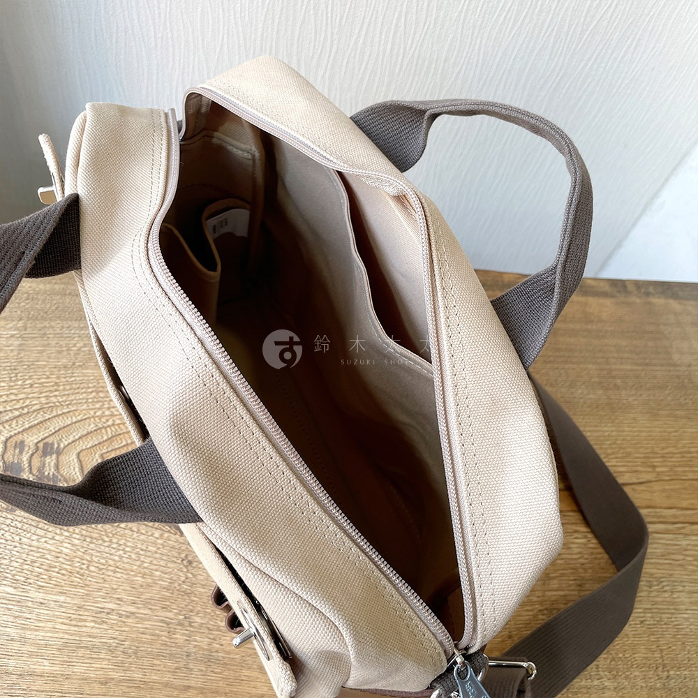 淺卡其色純棉帆布製成的橫式側背包，打開主隔層，露出內部多個隔層。背包帶為深棕色。