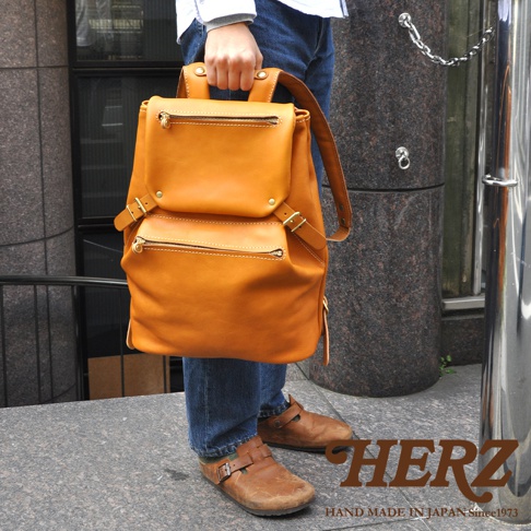 職人手作皮革多口袋後背包 R-135，展現溫潤的駝色皮革質感，設有雙層拉鍊前袋及金屬釦環，搭配藍色牛仔褲與棕色皮鞋，整體風格簡約實用。