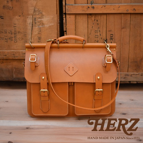 職人手作A-102皮革旅行包，採用經典棕色皮革，配有金色金屬釦件和可調節肩帶。包款設有頂部提把、翻蓋設計及雙前袋，縫線精緻，展現皮革的自然紋理。