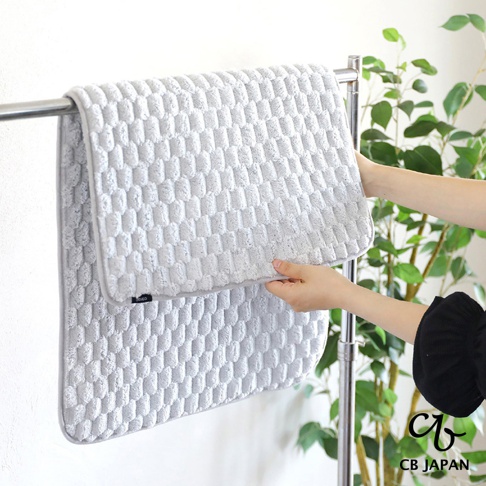 淺灰色格紋地墊懸掛在毛巾架上，兩隻手正在展示其中一塊地墊，背景有綠葉植物。