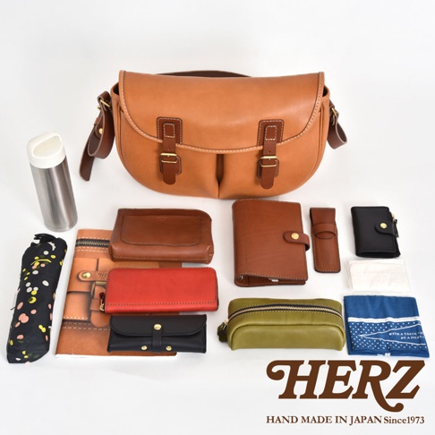 職人手作皮革傳統釣魚包，主色為棕褐色。包款為翻蓋式設計，配有兩個皮質搭扣。周圍展示多種配件，包含皮質筆記本、錢包、零錢包、雨傘及保溫瓶等，材質多為皮革，部分配件顏色有紅、黑、綠。