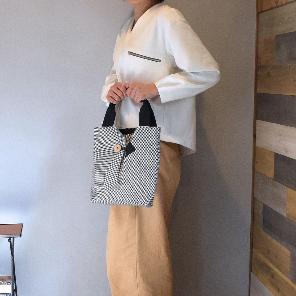 一個直立式托特包，採用黑白細小格紋的刺子織布料製成，配有黑色提把。包包前方有一個木製鈕扣，用於固定一個三角形的翻蓋。