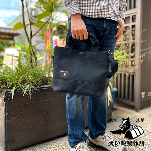 黑色帆布兩用托特包，方形包身，頂部有提把和旋轉釦開合，側面有可拆卸斜背帶。包包正面有一個帶有日文標誌的織標。