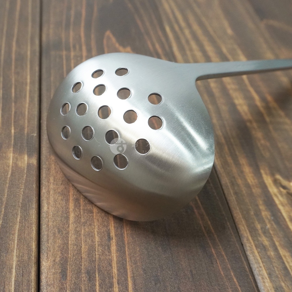 一個銀色不鏽鋼萬用瀝水湯勺，勺面有規則的圓形漏孔，勺柄為細長設計，放置在木紋背景上。
