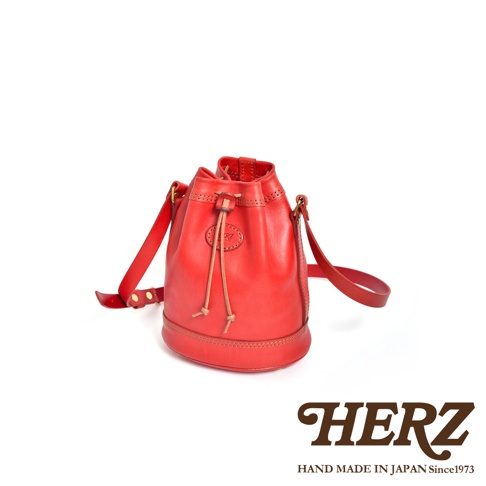 紅色職人手作皮革束口單肩包，附有可調節的皮革背帶和抽繩閉合設計。包包呈現圓潤桶狀，底部拼接皮革，質感紮實。