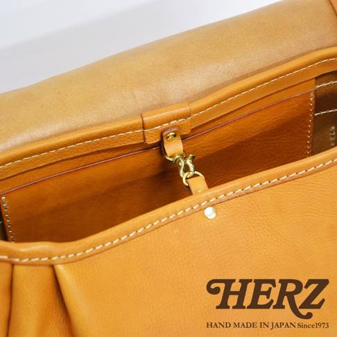 焦糖色皮革復古翻蓋郵差包內部，可見縫線、金屬鉤扣及品牌標誌。