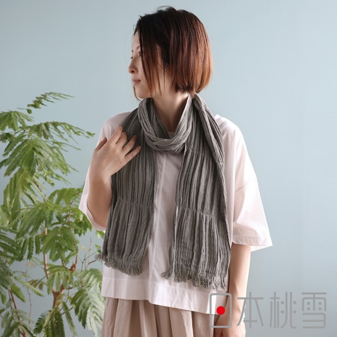 一位短髮女性模特兒穿著白色上衣與米色裙裝，脖子上圍著一條具有細膩皺褶紋理的灰色純棉紗布圍巾，圍巾兩端呈現自然的流蘇邊設計，整體風格簡約清新，背景為淺藍色牆面與一株綠色盆栽，右下角印有日本桃雪品牌識別文字。