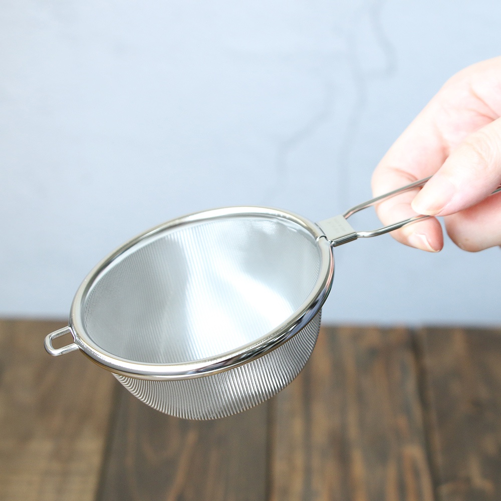 一只手持著不鏽鋼濾茶器，濾網為超細目，呈現銀色金屬光澤，帶有簡潔的提柄。