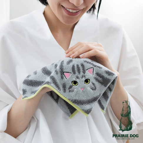 一位女性微笑著雙手捧起一條 Prairie Dog 品牌出品的灰色貓咪圖案刺繡方巾，方巾上有精緻的貓咪臉部刺繡，並採用觸感柔軟的材質與亮色滾邊設計，畫面呈現居家生活質感。