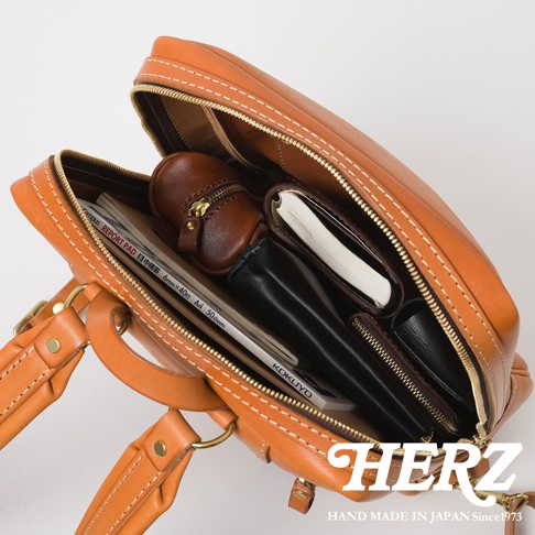 職人手作棕色皮革軟皮商務後背包，開口處可見內部收納物，包含筆記本、文件夾、零錢包，細節處縫線清晰，質感良好。