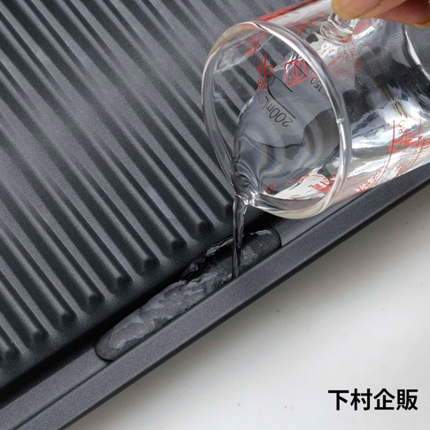 深灰色波浪紋烤盤，烤盤表面有條紋，一個透明玻璃量杯傾倒清水，水流進入烤盤的凹槽中。