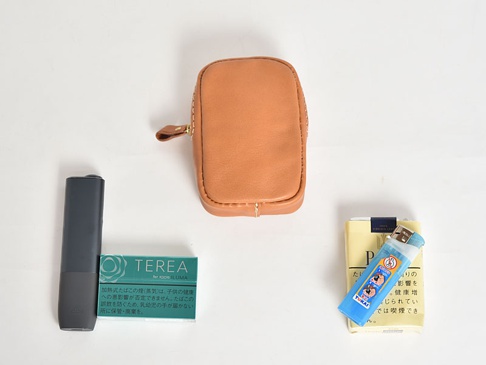 一個棕色皮革製成的職人手作收納包，帶有拉鍊和皮革拉帶。旁邊擺放著一個深灰色電子菸裝置、一盒綠色包裝的加熱式菸草，以及一盒黃色包裝的香菸，上面放著一個藍色打火機。