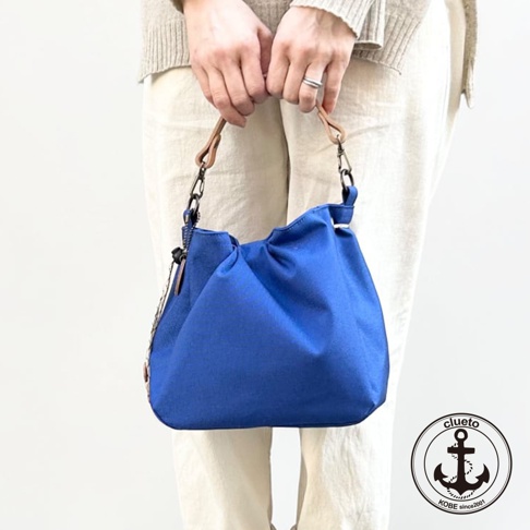 一個人雙手提著一只神戶clueto品牌旗下的BALON mini防潑水藍色手提包，包款採用簡約的立體剪裁設計，搭配淺棕色皮質提把與金屬扣環，模特兒身穿米色針織衫與亞麻長褲，畫面右下角帶有clueto的船錨標誌。