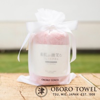 粉紅色潤澤素肌微笑速吸毛巾，以白色半透明紗網袋包裝，並繫上白色緞帶蝴蝶結。毛巾上印有日文「素肌が微笑む」及英文「bare skin smiles」字樣。