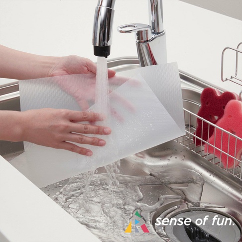 一雙手正在水槽中沖洗一張半透明、帶有網格紋路的白色捲筒式砧板保潔墊片，水流從水龍頭傾瀉而下，墊片上有水珠。