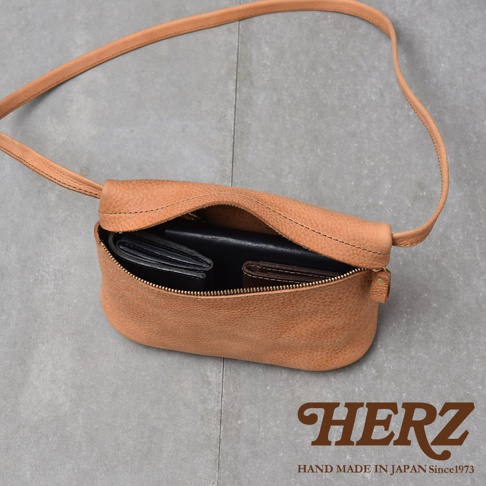 駝色皮革製作的斜背包，包內裝有皮夾，包身旁壓印有HERZ字樣及HAND MADE IN JAPAN Since1973字樣