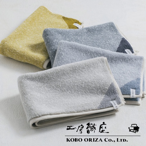 四條棉麻混紡浴巾，顏色為芥末黃、淺灰、藍灰及深藍，浴巾邊緣有細緻的包邊，部分浴巾角落帶有雙色拼接設計。